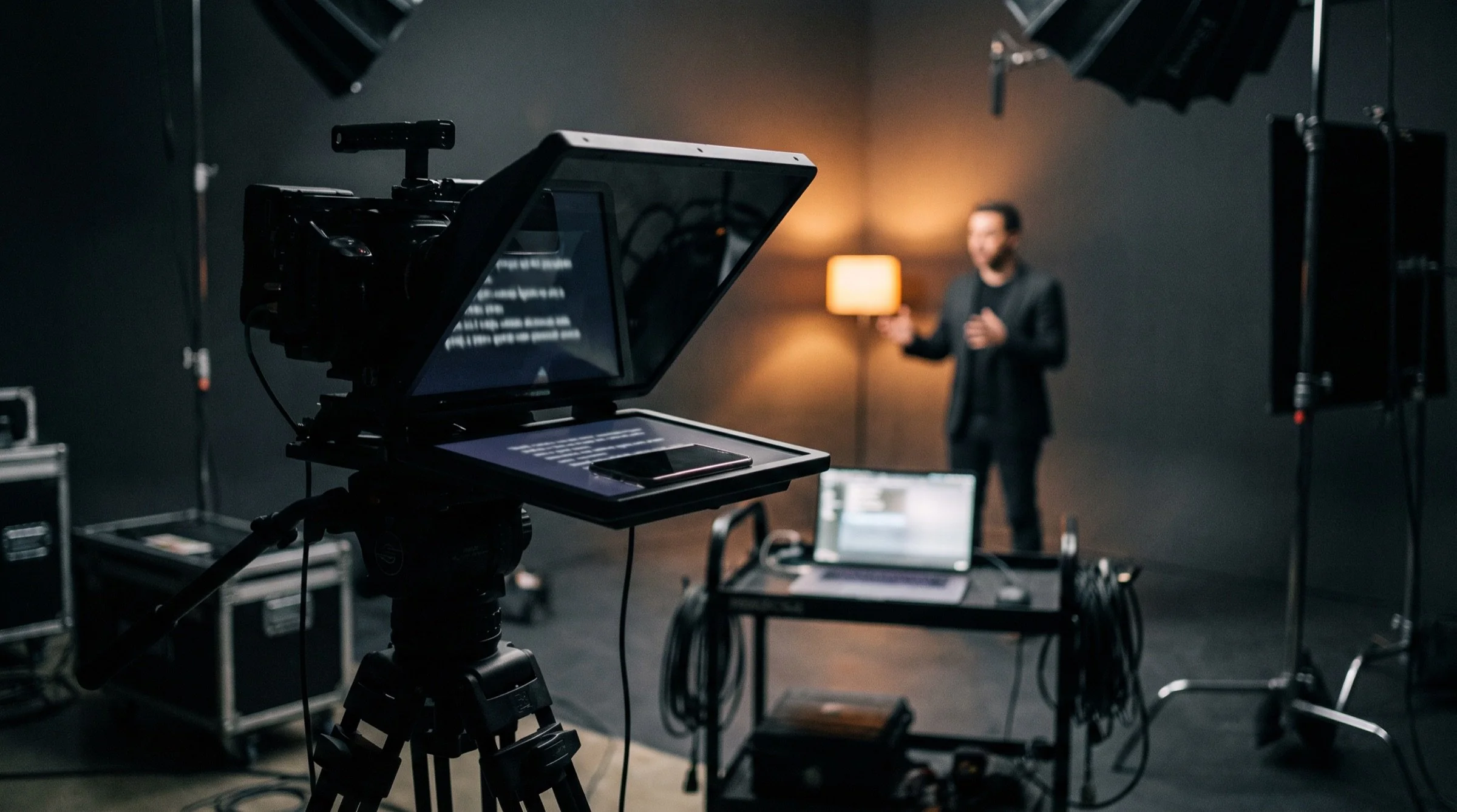 A cinema camera with a teleprompter hood on set, a phone in the hood, a laptop on a cart and a presenter in warm light behind
