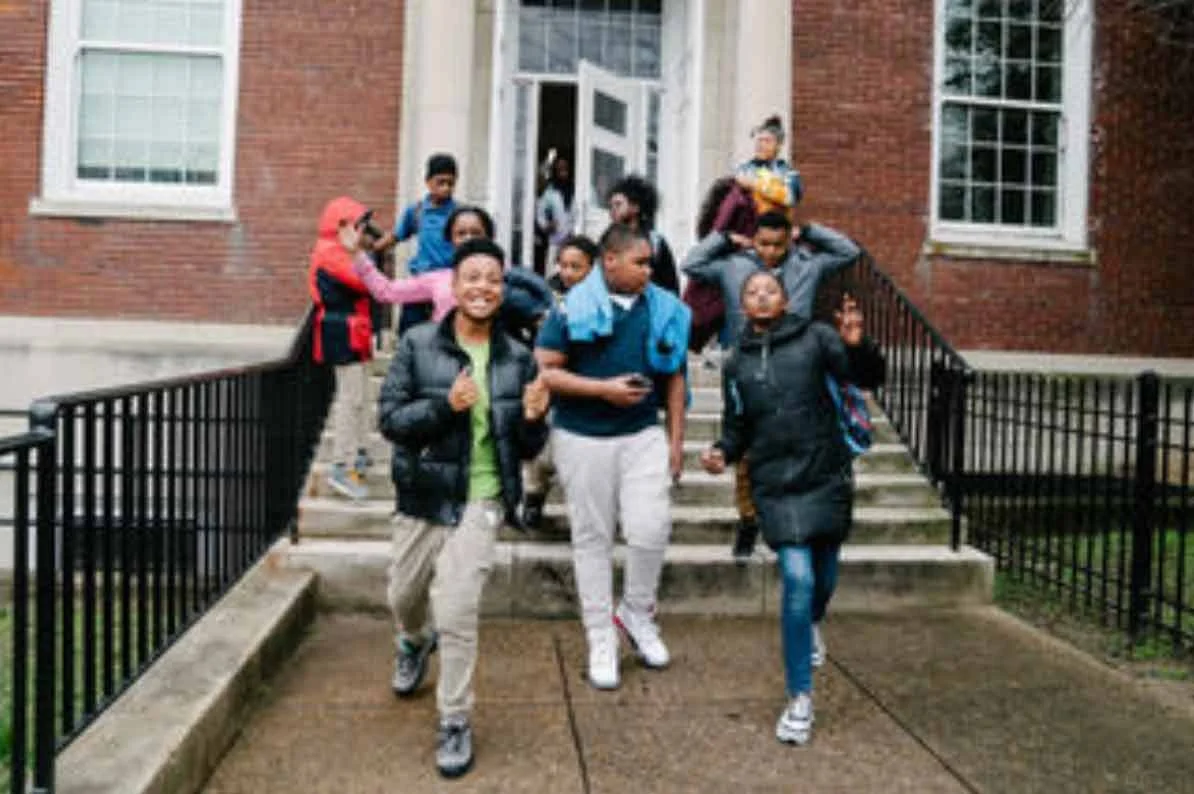A group of teens exits down the front stairs of their school smiling.