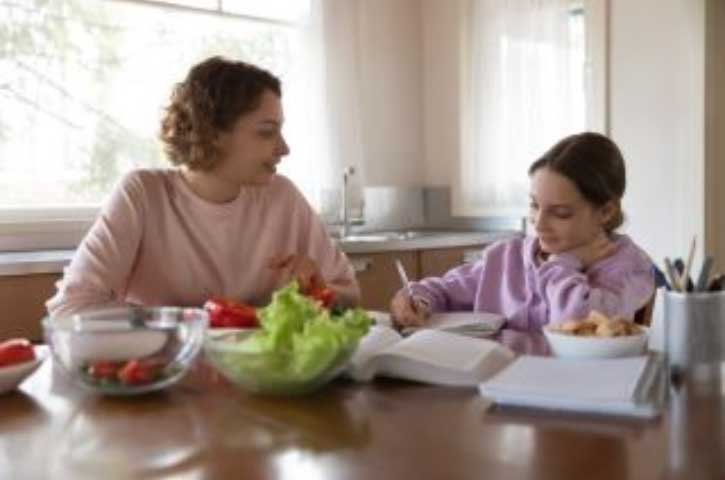 A mother sits next to her child at a kitchen table while the child works on a homework assignment.