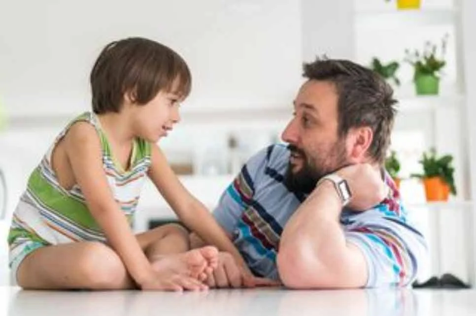 A young boy sits on a table in conversation with his father.