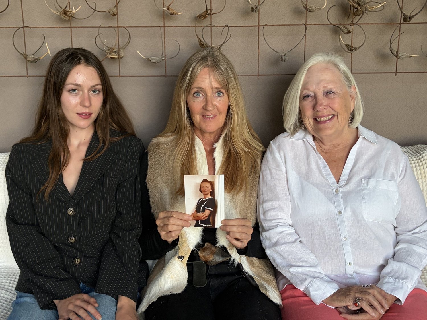 Olivia, Christie and Marsha Ruth on a sofa beneath a wall of antlers; Christie holds a photograph of her grandmother, Irma.