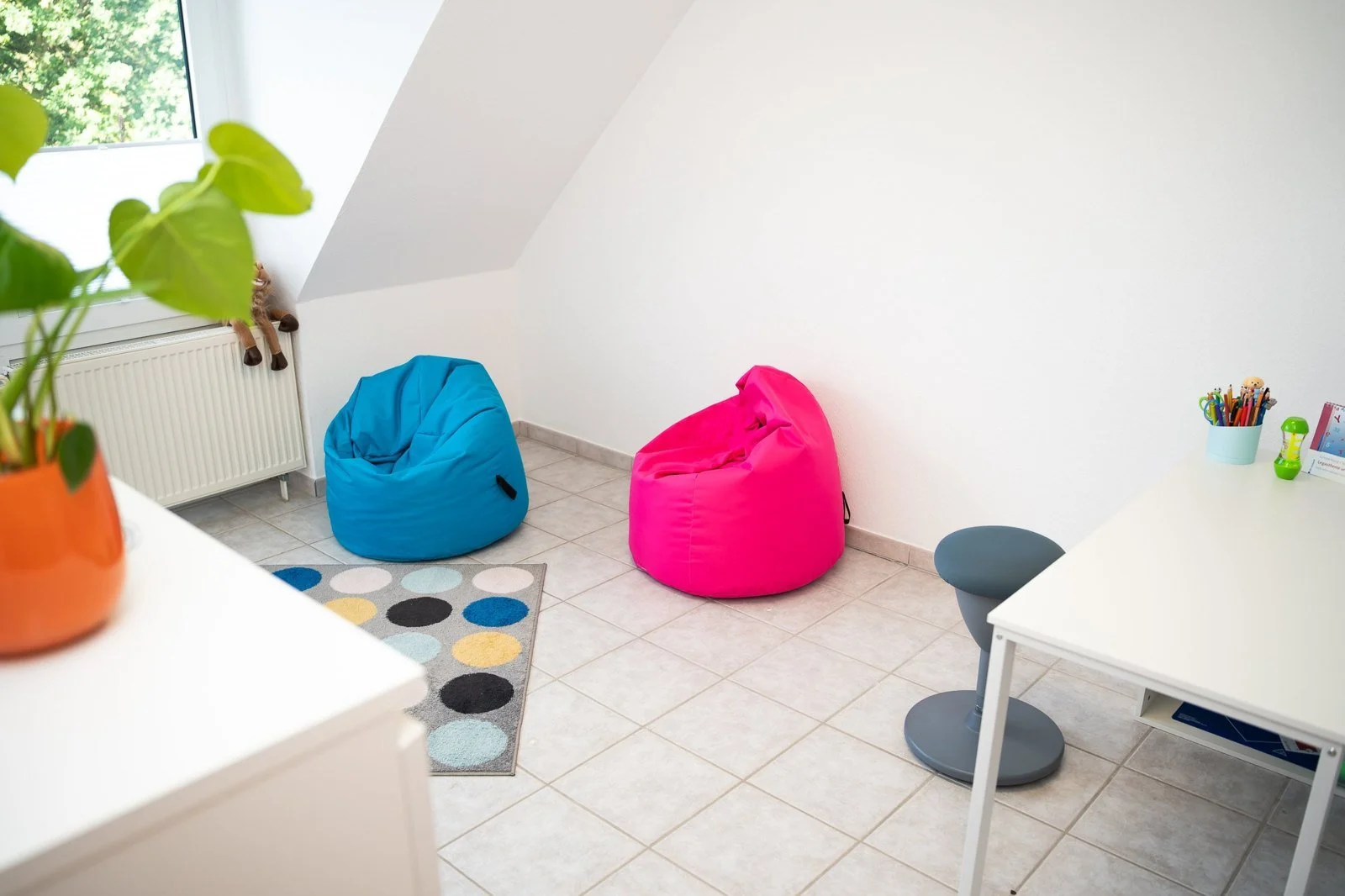 Heller Therapieraum mit Sitzsäcken in Blau und Pink, gepunktetem Teppich und Arbeitstisch
