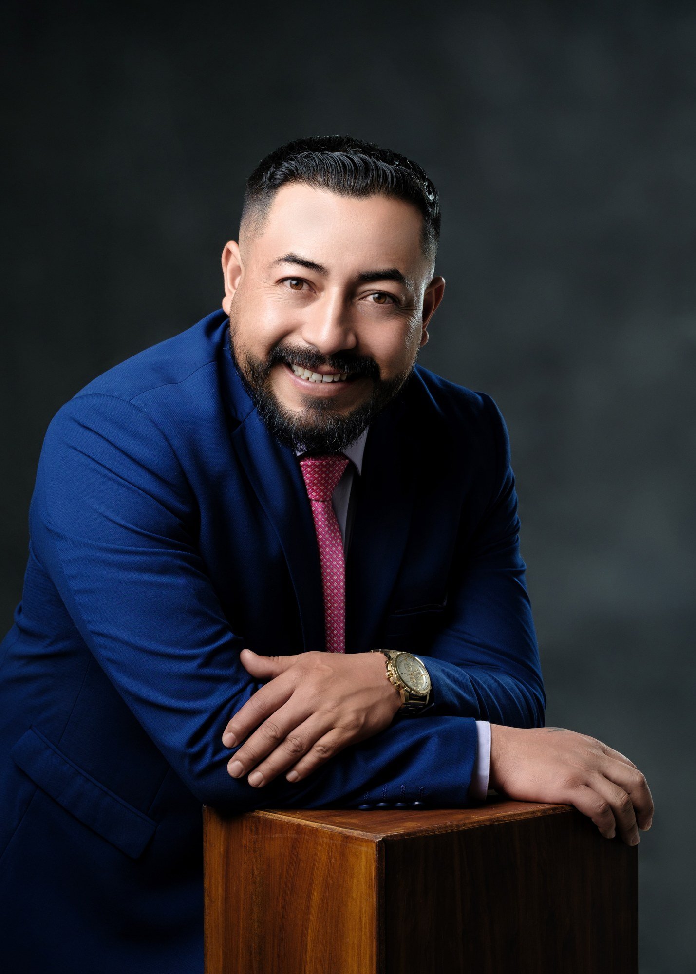 Hombre con traje azul sonriendo y apoyado en un soporte de madera.