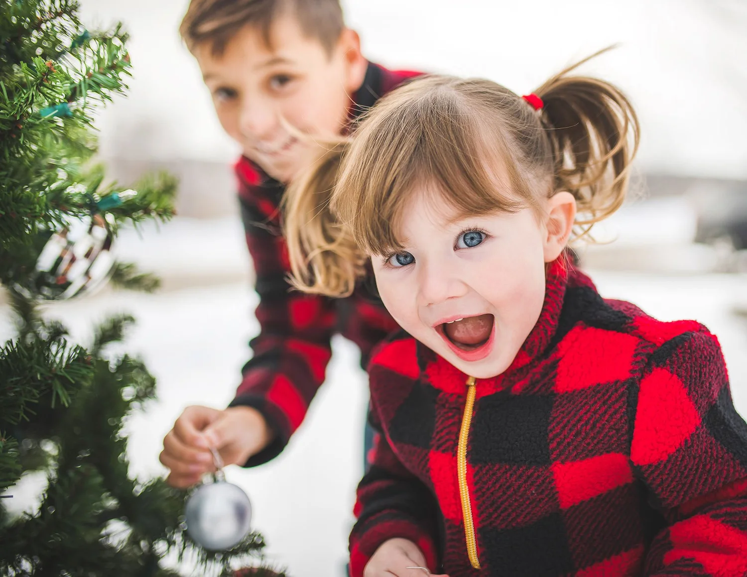 Two children in red plaid smiling beside a Christmas tree