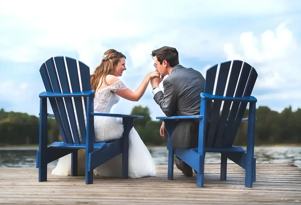 Groom kissing the bride’s hand as they sit in blue Muskoka chairs on a lakeside dock