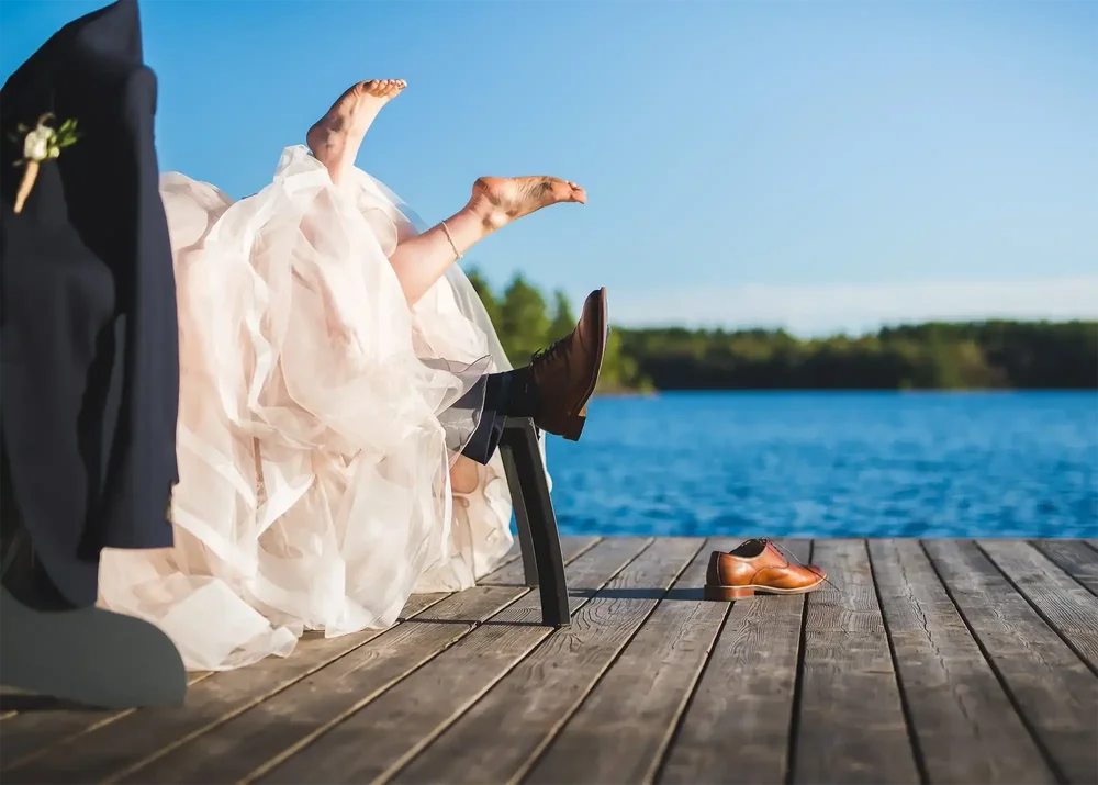 Playful wedding photo with the couple’s feet in the air behind Muskoka chairs on a dock