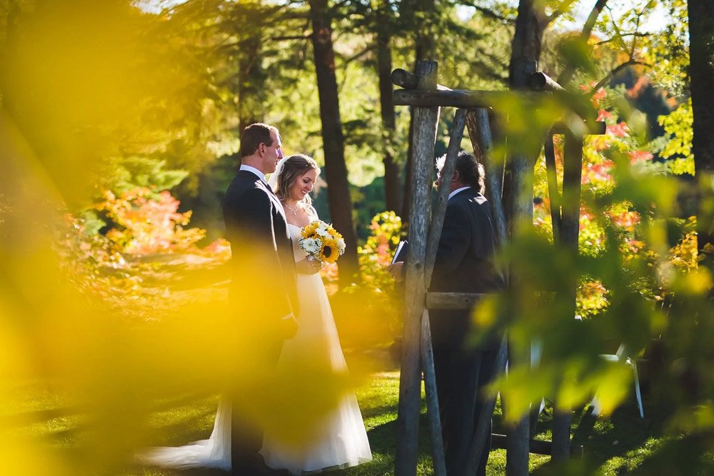 Couple exchanging vows beneath a wooden arbour surrounded by golden autumn leaves