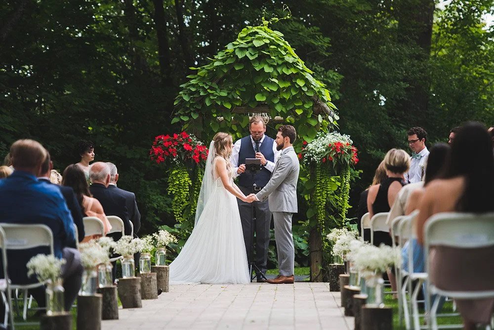 Bride and groom exchanging vows beneath a leafy arbour during an outdoor wedding ceremony