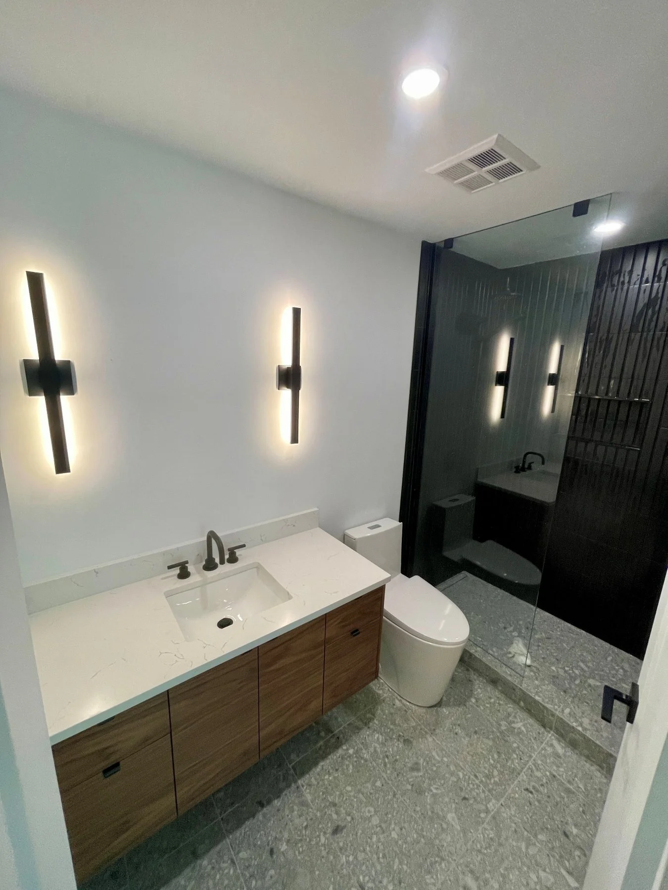 Distillery District condo bathroom after the renovation, with a walnut floating vanity, LED sconces and black-tile shower