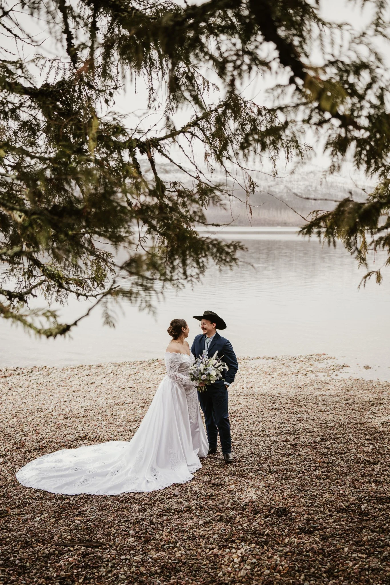 Ian and Rachel standing together beneath a cedar tree at the 7-Mile Pullout, smiling at each other, mountains and lake behind them