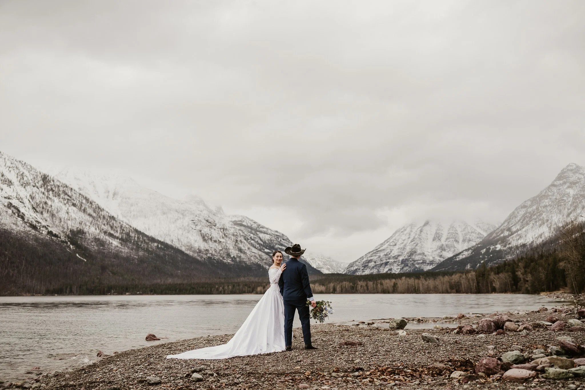 Ian and Rachel at Lake McDonald Lodge beach, snow-covered mountains reflected in the still water