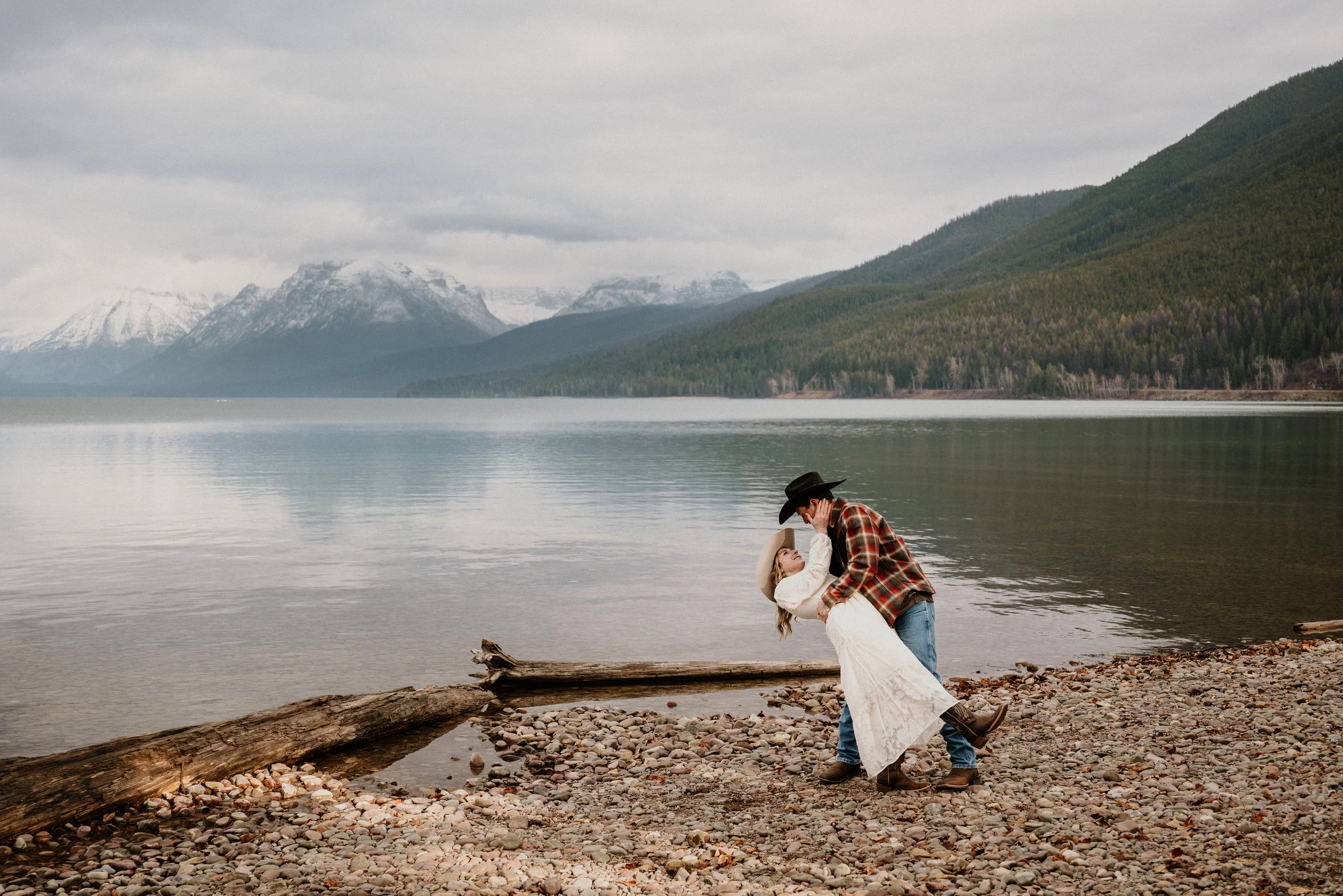 Dip kiss on the rocky shore of Lake McDonald with snowy Glacier National Park peaks in the background