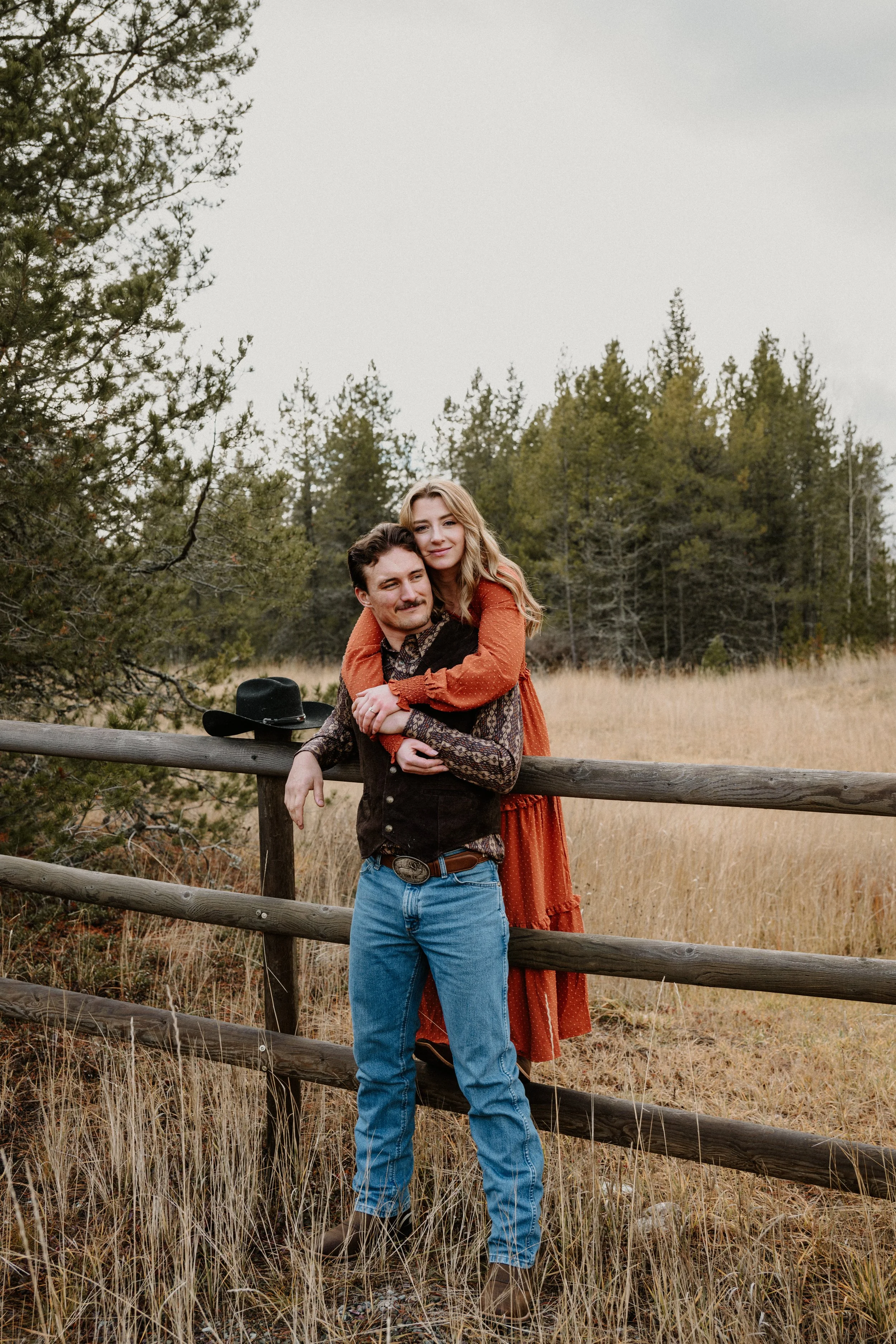 Engagement session at Belton Stage Park in West Glacier, Montana — couple leaning on a ranch fence with a black cowboy hat