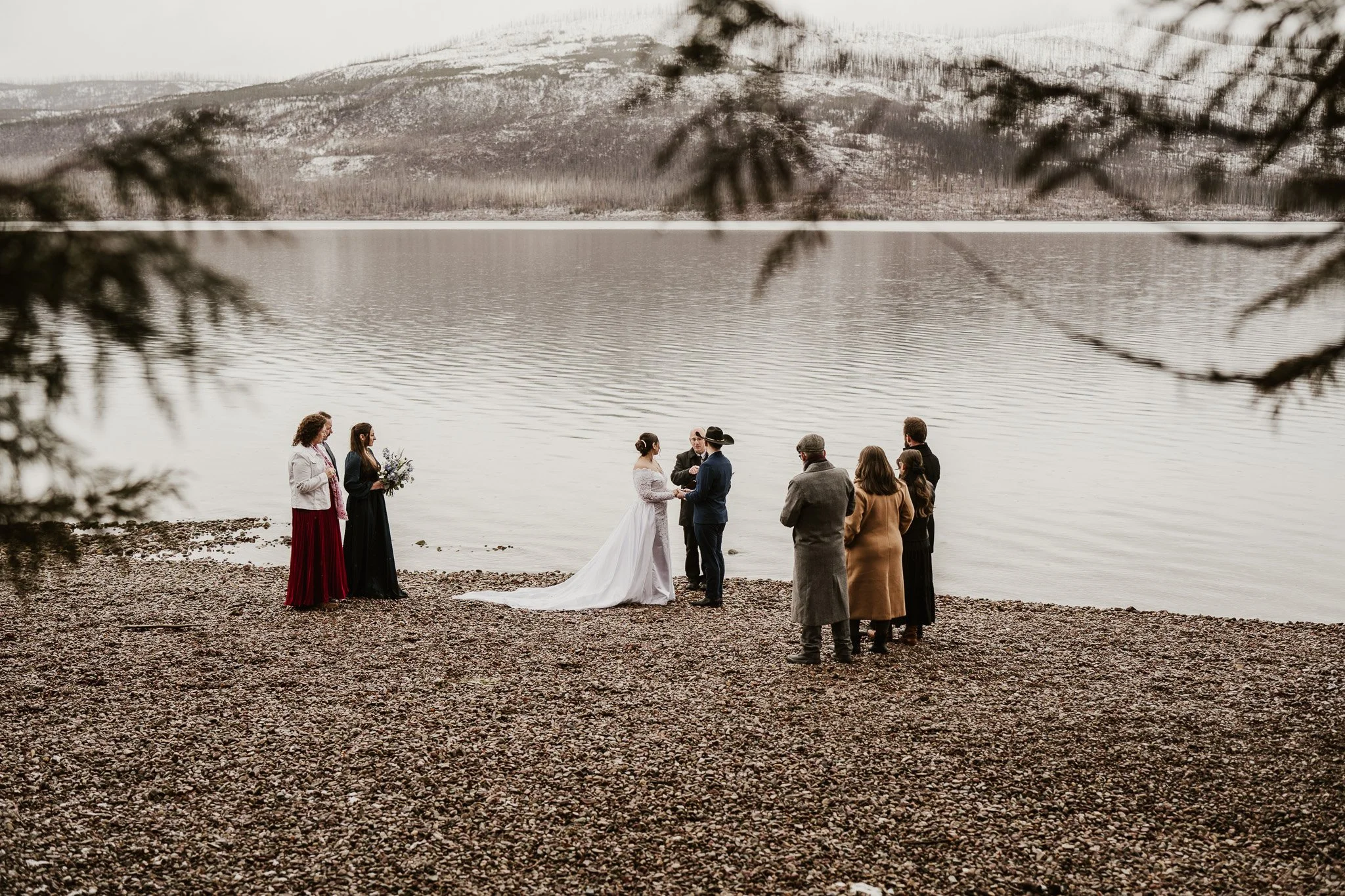 Ian and Rachel during their ceremony at the 7-Mile Pullout, Glacier National Park, family gathered around them on the pebbled shore