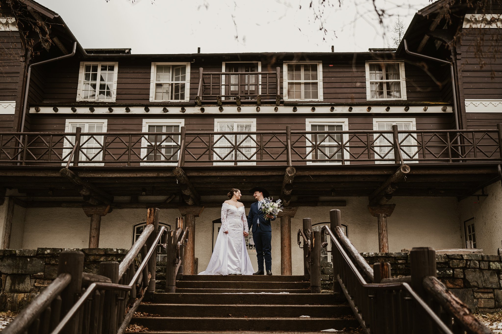 Ian and Rachel standing on the stone steps of Lake McDonald Lodge, holding hands and looking at each other, the historic timber facade behind them