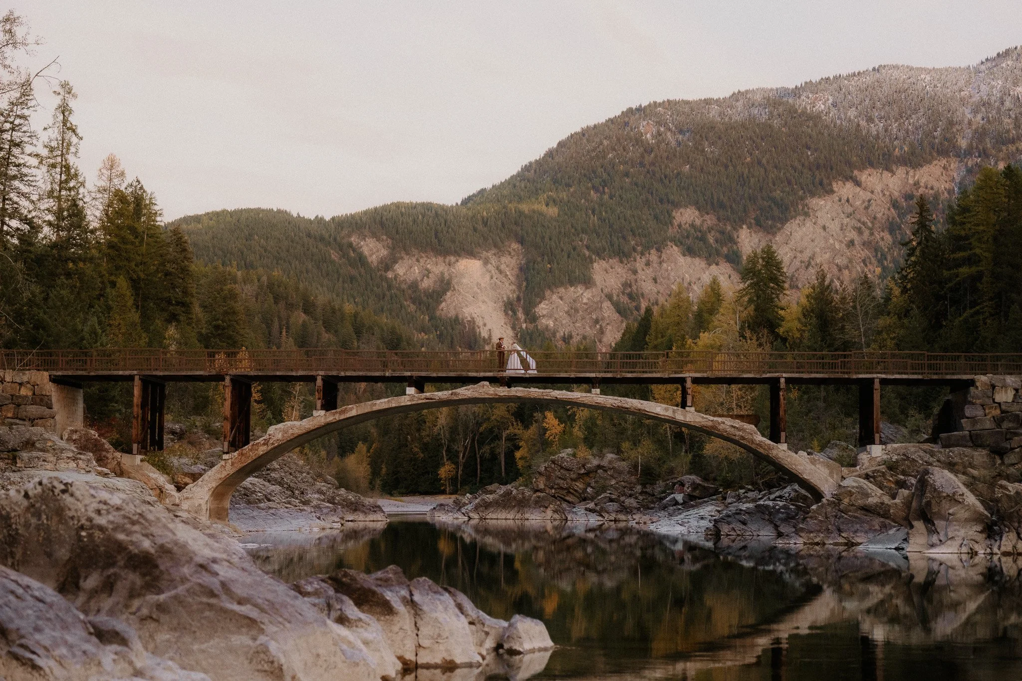 Wide view of a bride and groom on a historic stone arch bridge in Glacier National Park at golden hour