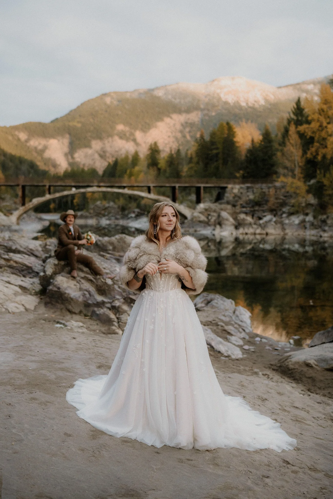 Bride in a fur shawl with her groom seated on a rock near a historic stone bridge in Glacier National Park