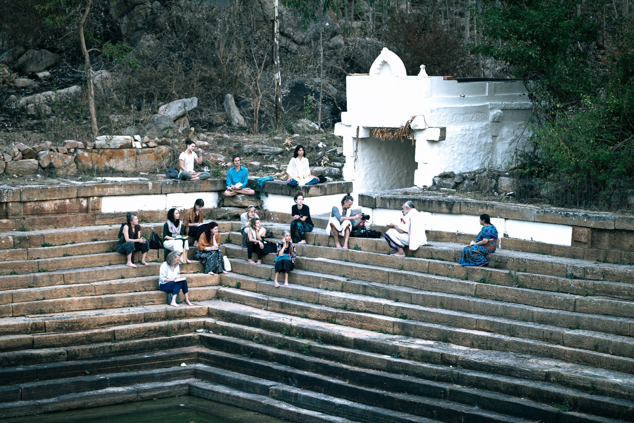 Foreign and Indian students gathered on the stone steps of a temple tank in Mysore, listening to a scholar teach, with a small camera setup on the steps recording the session