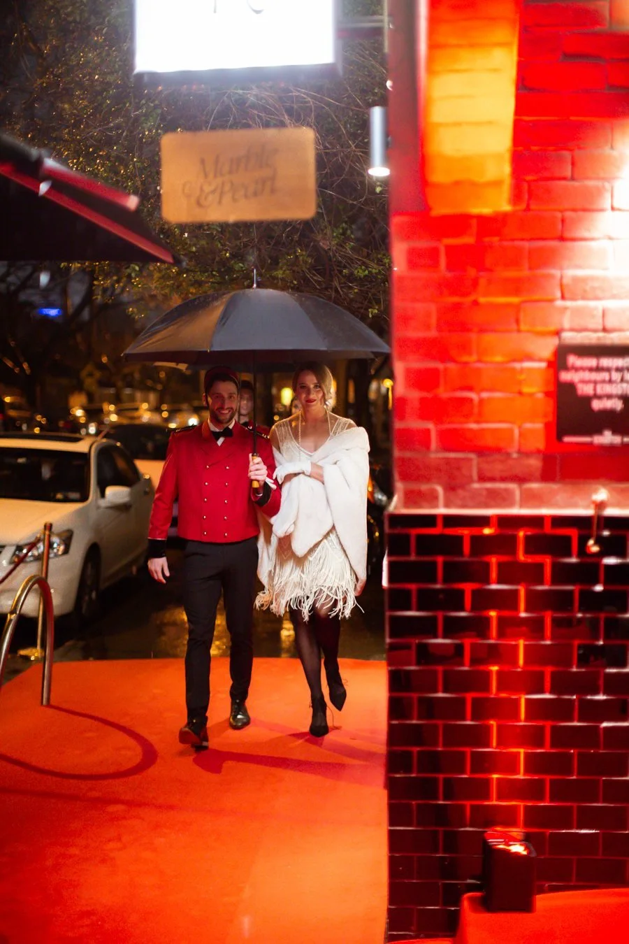 A red-coated bellhop escorts a guest under an umbrella on the red carpet