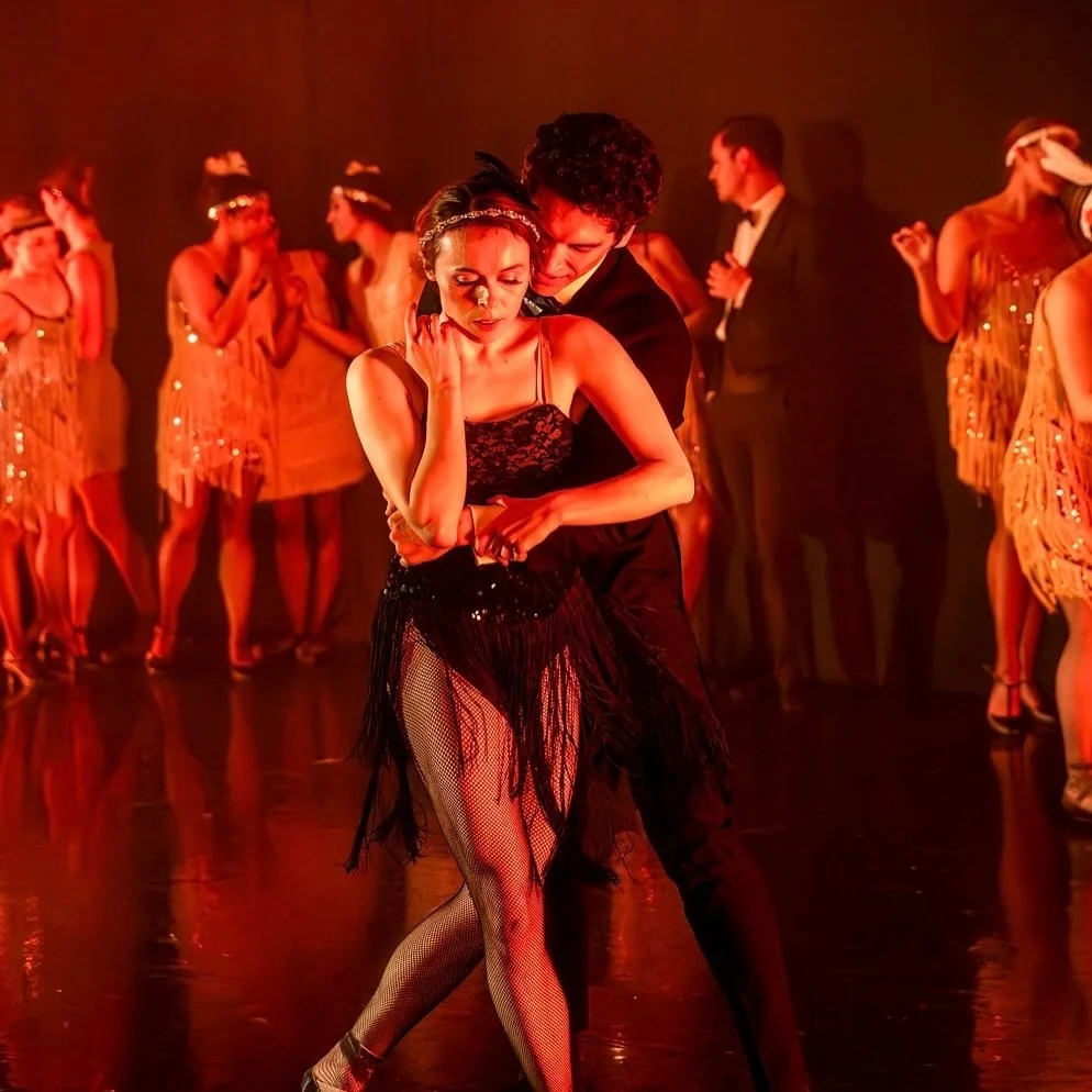 A couple in a close embrace, her eyes closed, flappers in sequined gold behind them.