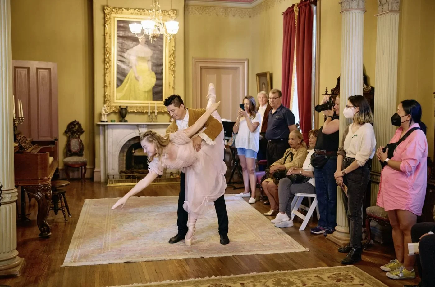 A ballerina in a pink gown is dipped by her partner in a Victorian parlor as guests watch up close.