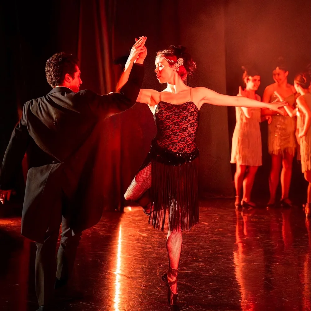 A dancer on pointe in a black fringed dress, arms extended, hand in hand with her partner.