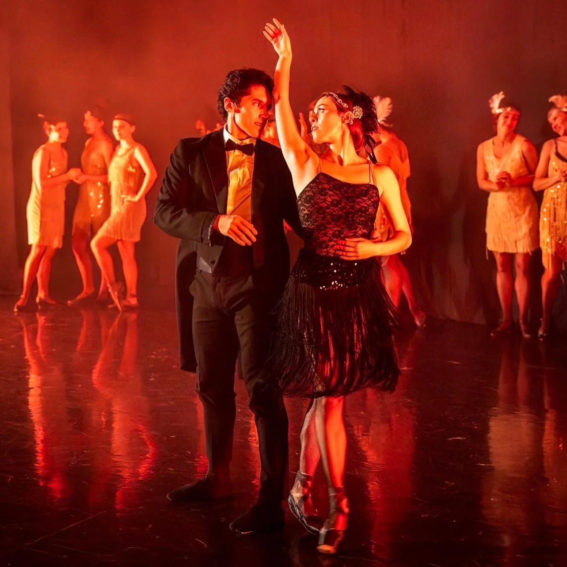 A dancer raises one arm high beside her partner in a tuxedo, flappers watching.