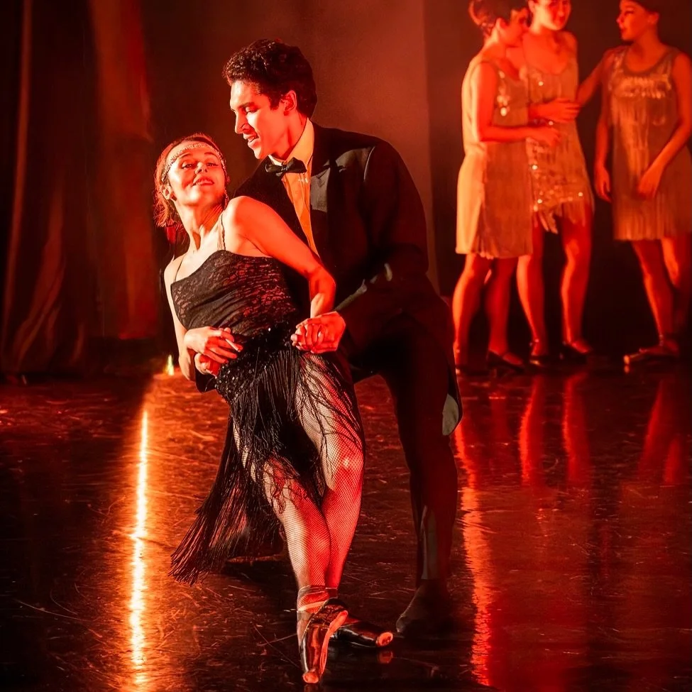 A dancer in a black fringed flapper dress is dipped by her partner in tails, with flappers in gold behind them under red stage light.