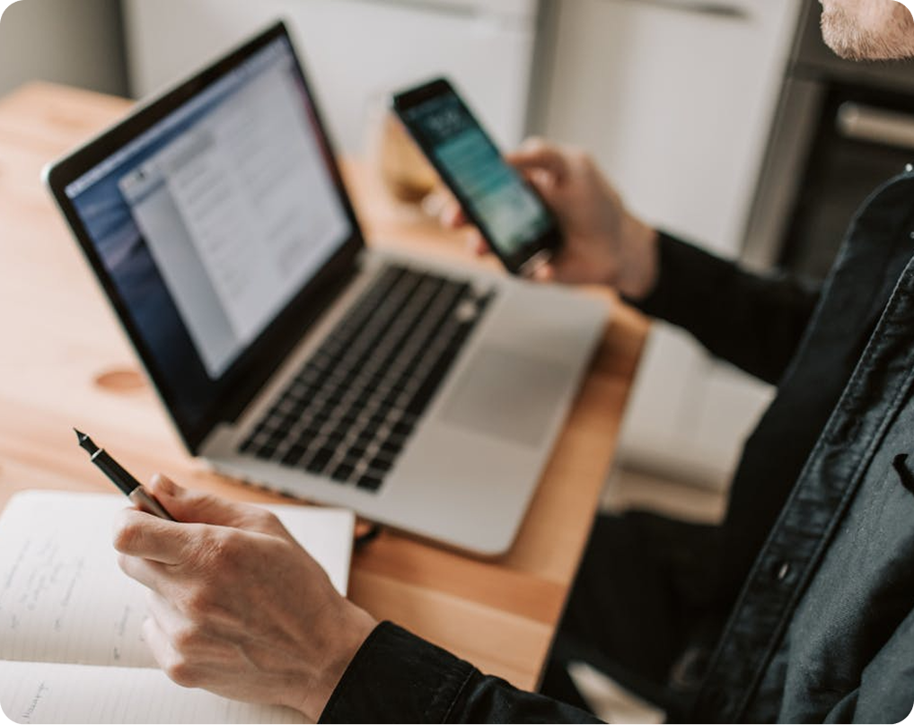 Someone sitting at a laptop, phone in one hand, pen in another