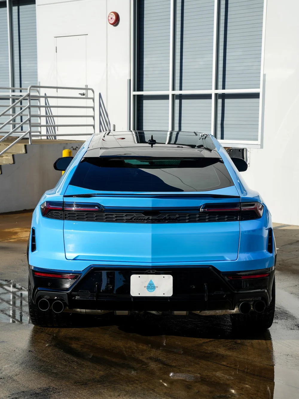 Centered rear view of the light-blue Urus SE with its rear light strip, black bumper and studio plate visible
