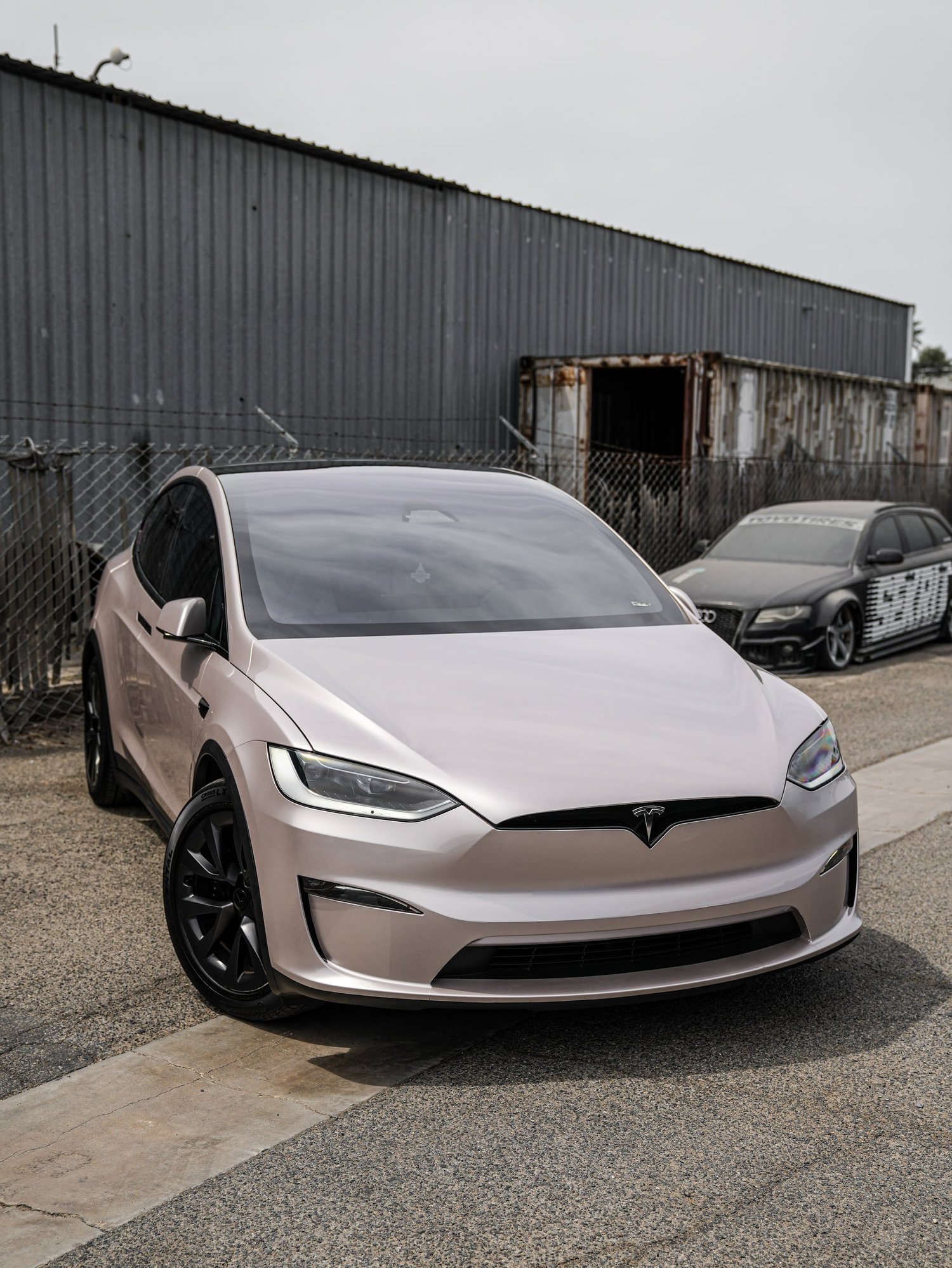 Complete front three-quarter view of the Champagne Gold Tesla Model X with dark wheels beside an industrial fence.