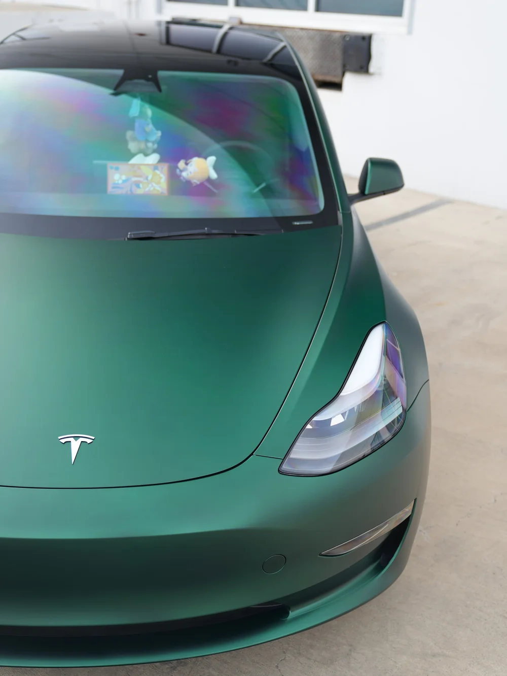 Tesla Model 3 hood and headlight beside Ghost Matte Green vinyl wrap in a close front view