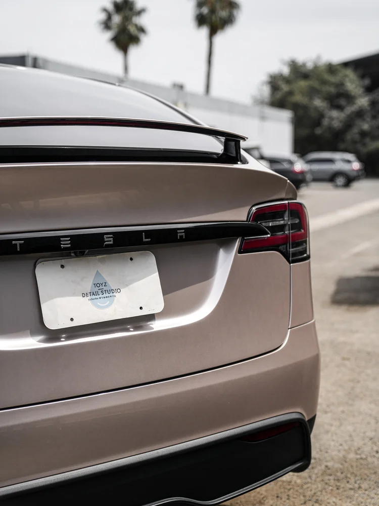Model X champagne-gold liftgate, spoiler and rear lamp beside a Toyz Detail Studio plate insert.