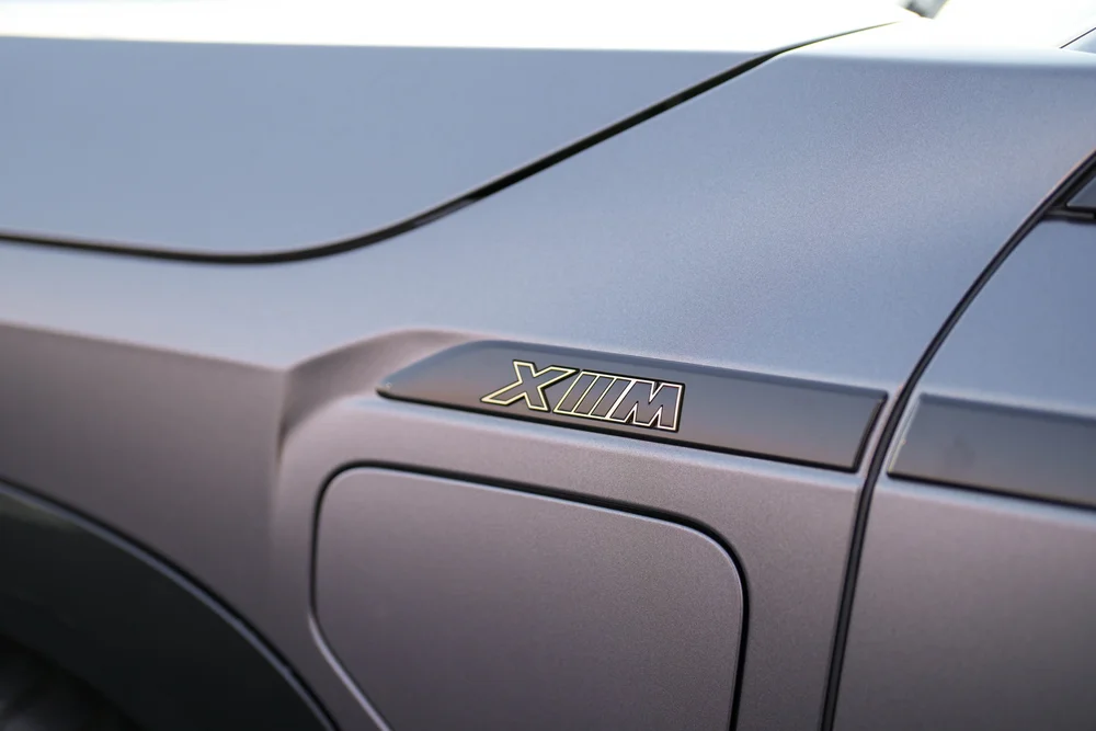 Close front-fender detail showing XM lettering above a rectangular flap, beneath the seam of the matte-grey hood.