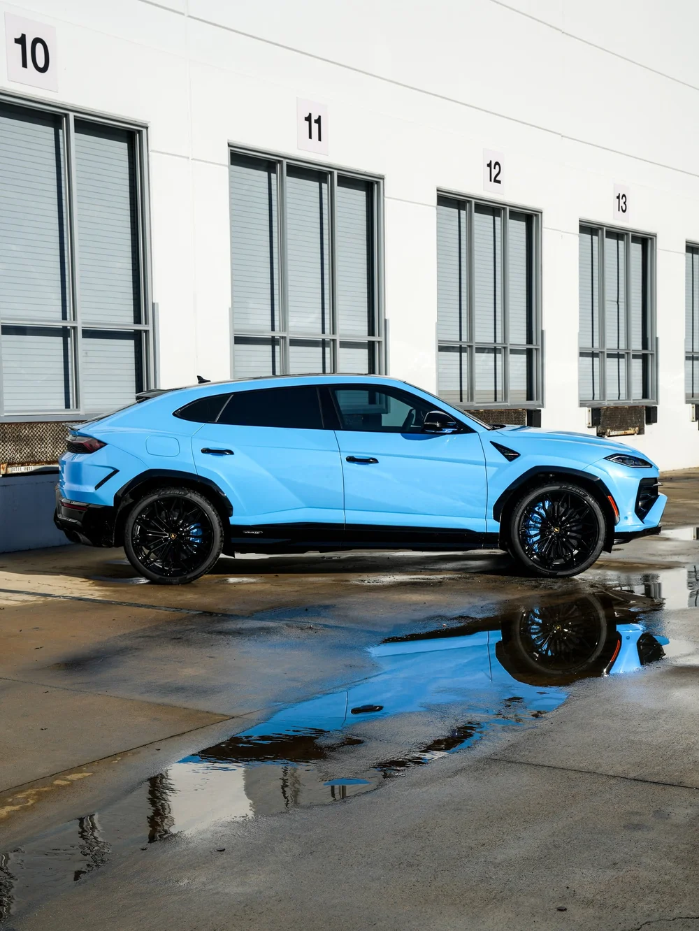 Full side profile of the light-blue Urus SE with dark side glass, black wheels and a reflection in a puddle
