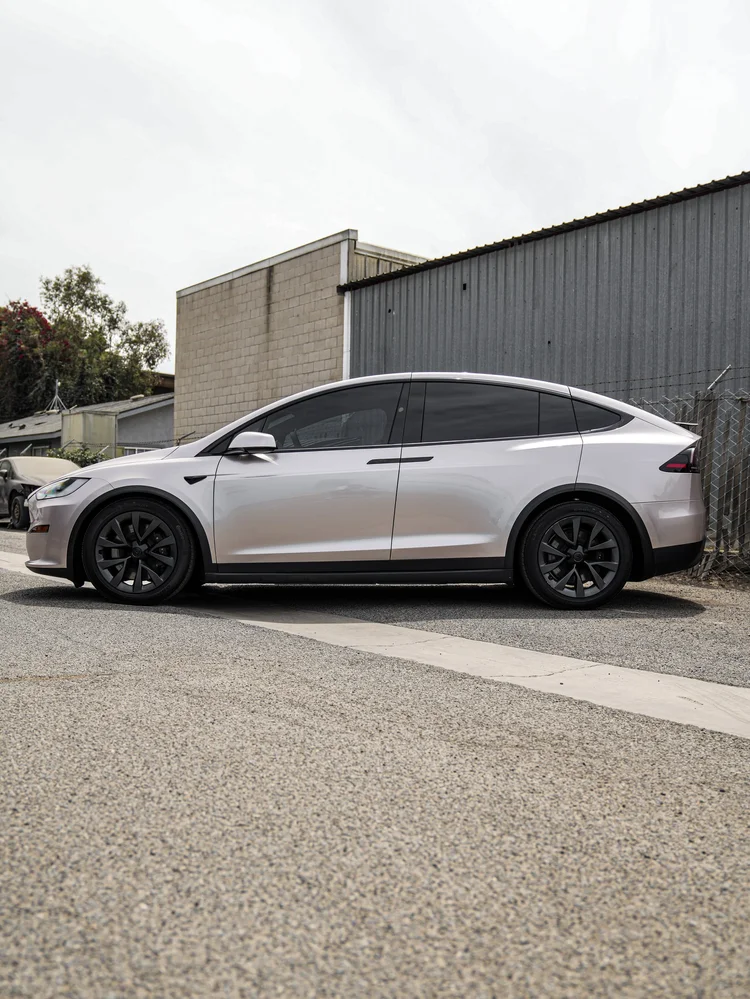 Complete side profile of the Champagne Gold Model X with dark glazing and wheels beneath an industrial building and sky.