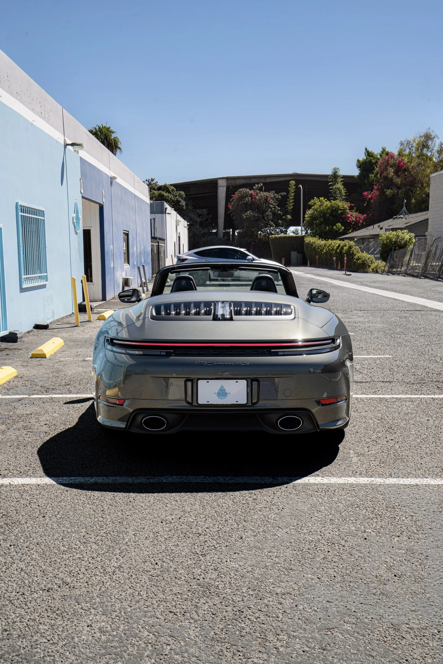 Straight-on rear view of the roof-down Vanadium Grey Porsche Cabriolet in the parking area beside Toyz Detail Studio.