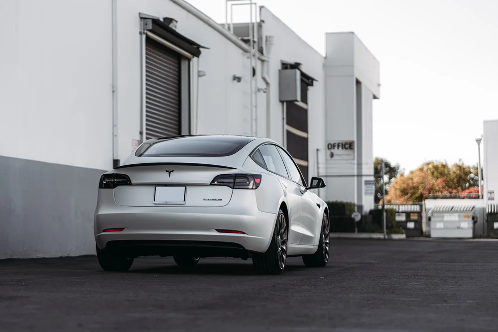 Wide low rear three-quarter view of the Tesla Model 3 with 3M 2080 Satin Pearl White vinyl and a dark rear spoiler