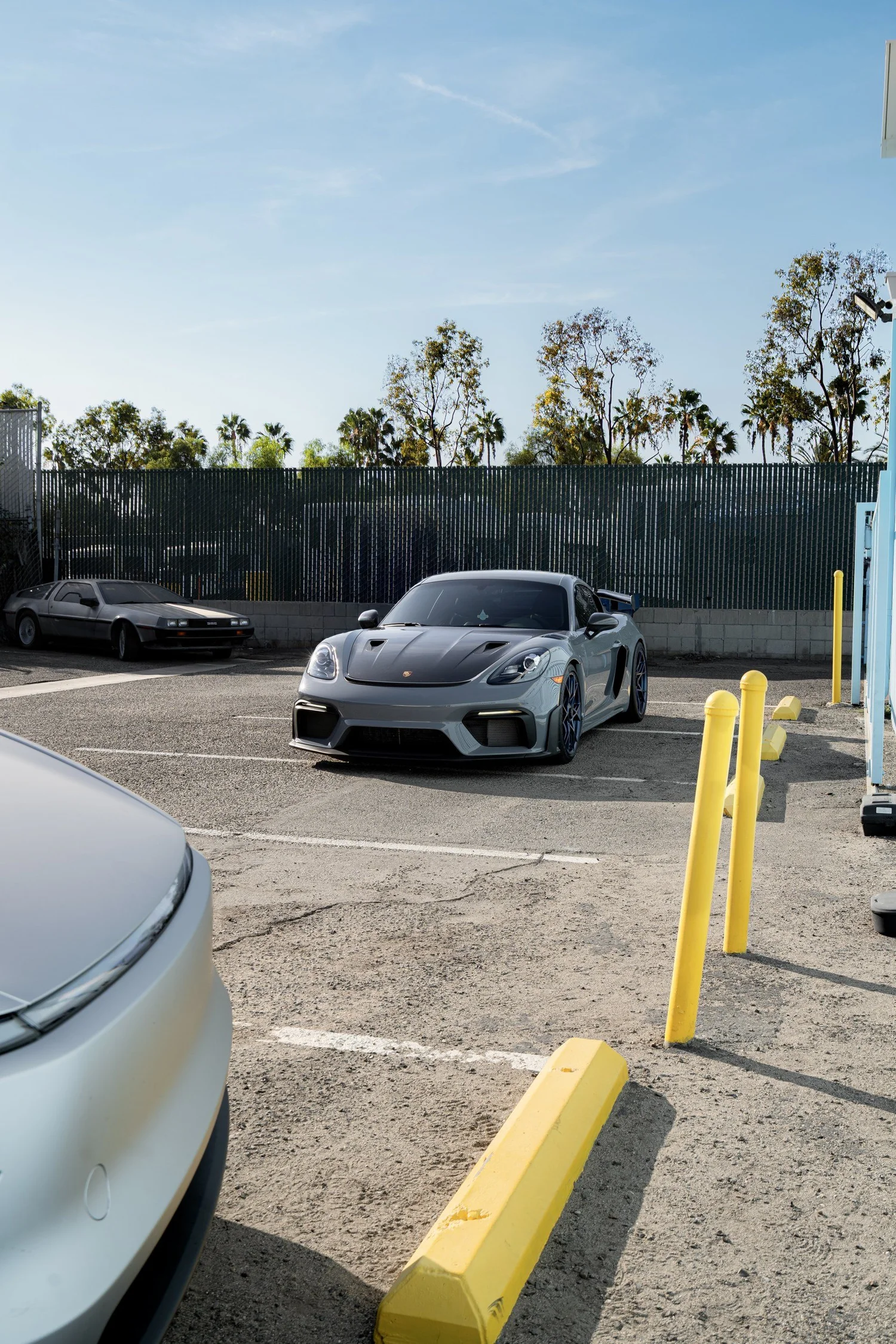 Distant front three-quarter view of the Arctic Grey GT4 RS in the Toyz yard, framed by yellow bollards and other parked cars.