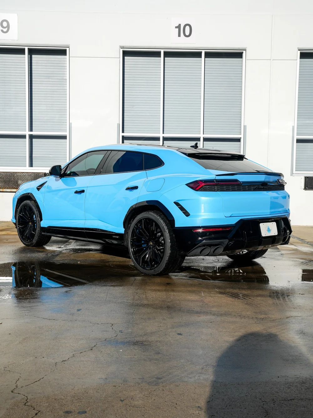 Full rear three-quarter view of the light-blue Urus SE with black wheels, dark side windows and twin exhaust outlets visible