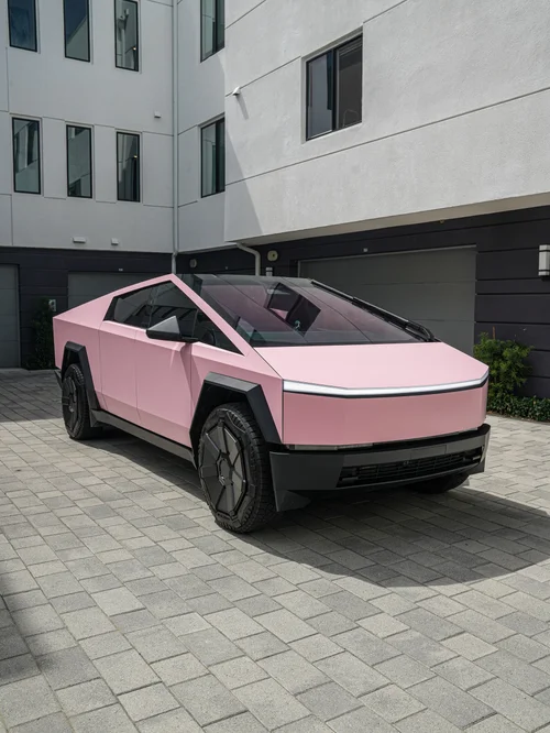 Pink Tesla Cybertruck in a front three-quarter view on paving beside residential buildings.