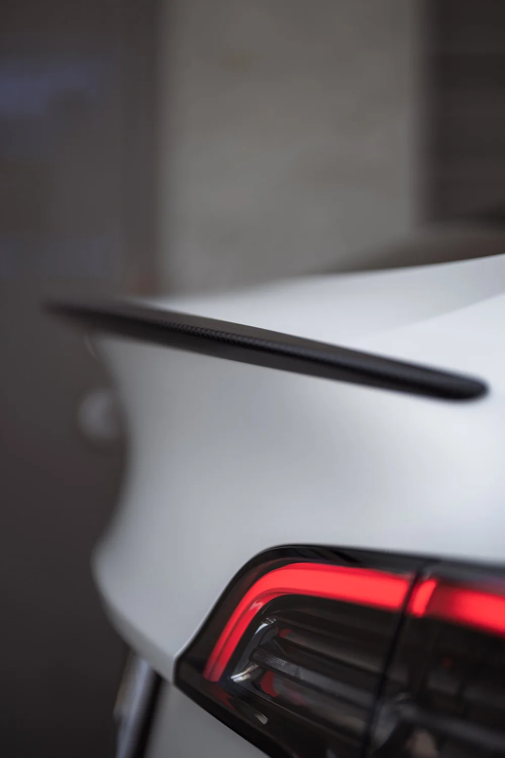 Close view of the dark rear spoiler above the Satin Pearl White Model Y hatch and illuminated taillight