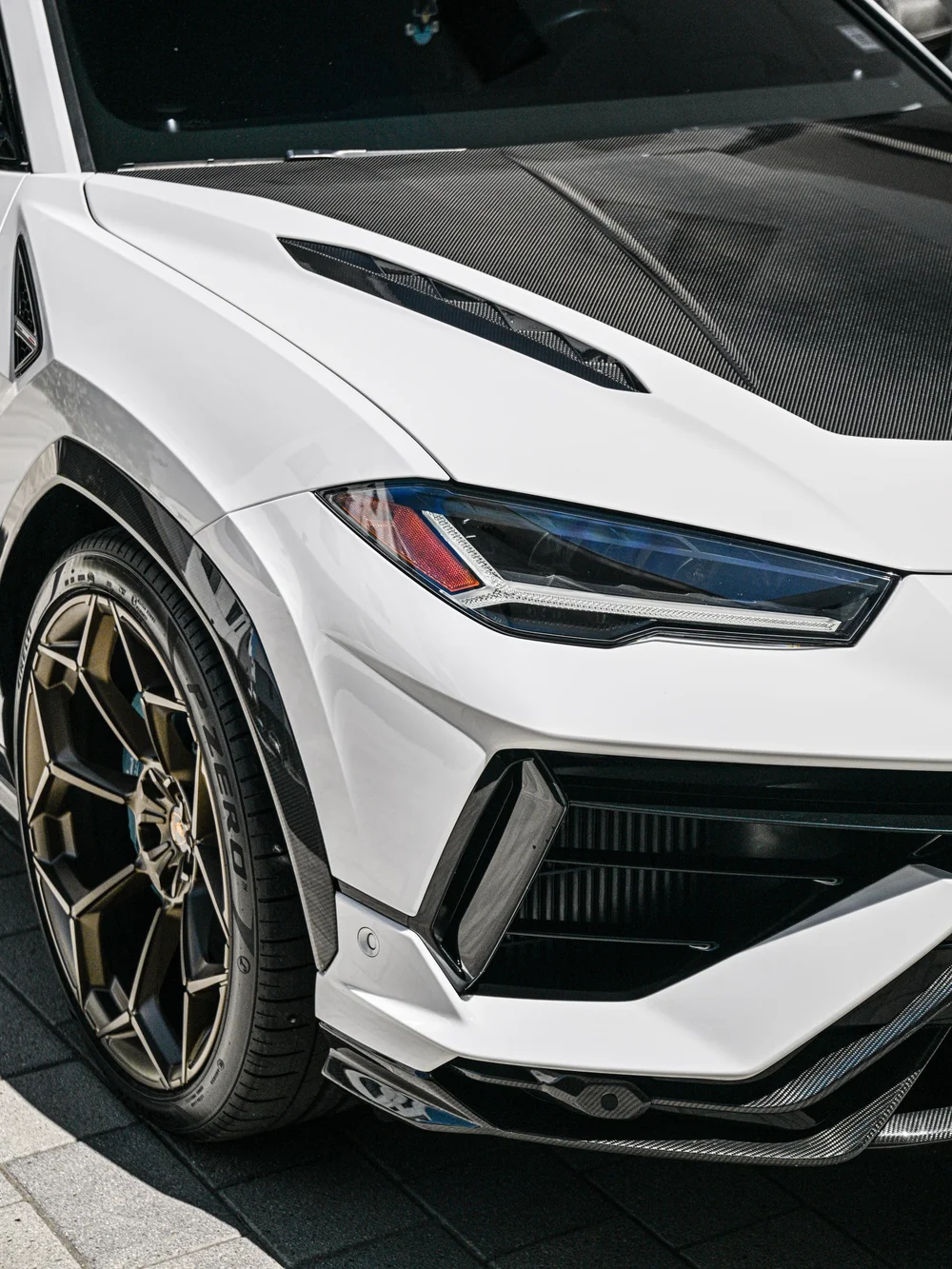 Urus headlight, white front bumper and bronze-toned wheel beside the dark hood