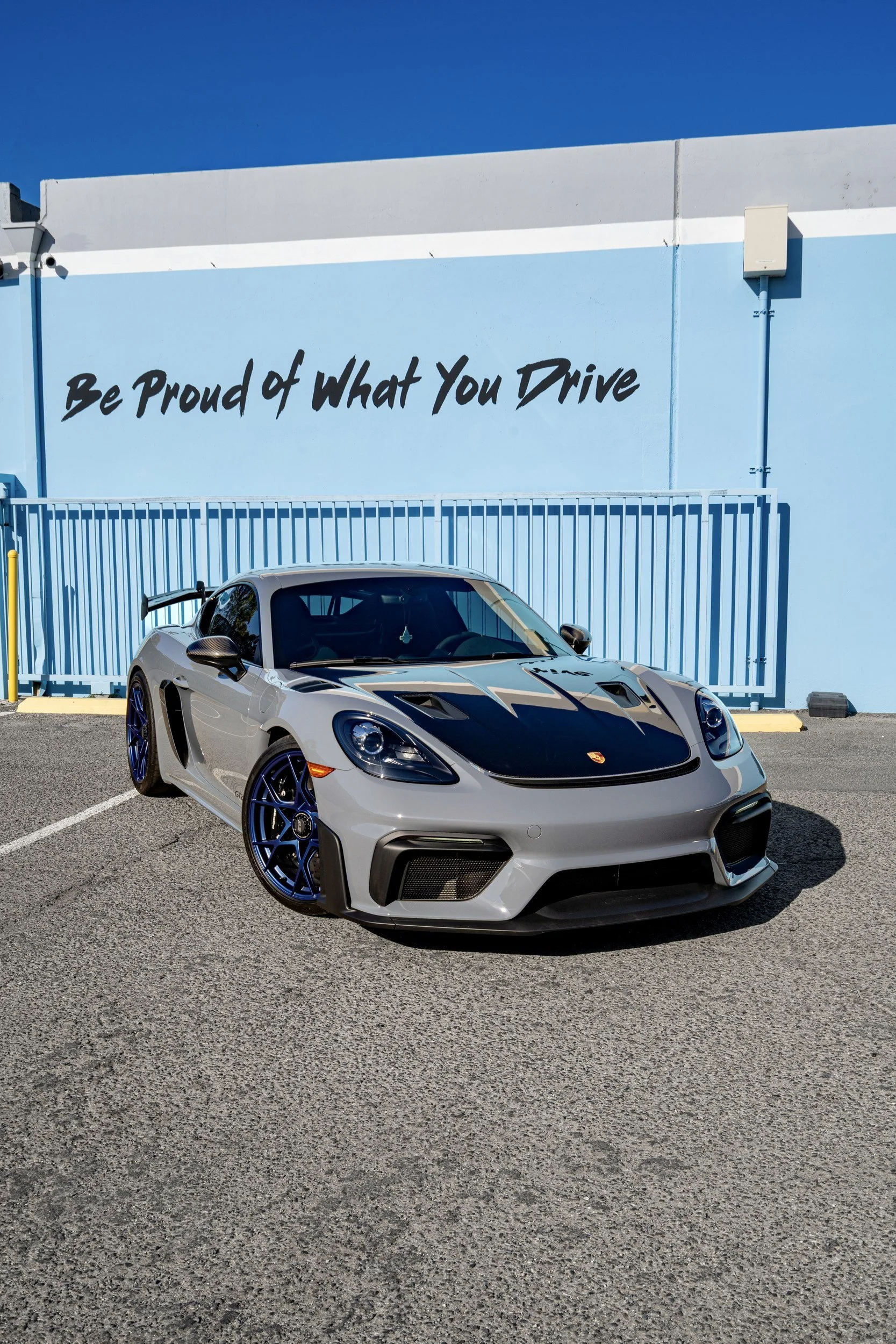 Front three-quarter view of the Arctic Grey 2025 Porsche 718 Cayman GT4 RS with exposed carbon hood and blue wheels outside Toyz Detail Studio.