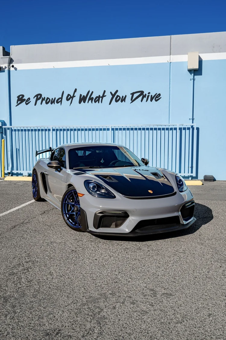 Front three-quarter view of the Arctic Grey 2025 Porsche 718 Cayman GT4 RS with exposed carbon hood and blue wheels outside Toyz Detail Studio.