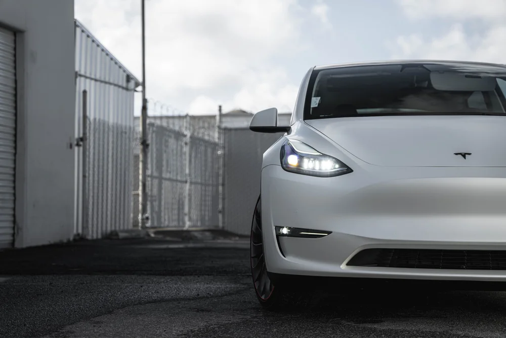 Wide front view of the Tesla Model Y showing the illuminated headlight and Satin Pearl White hood