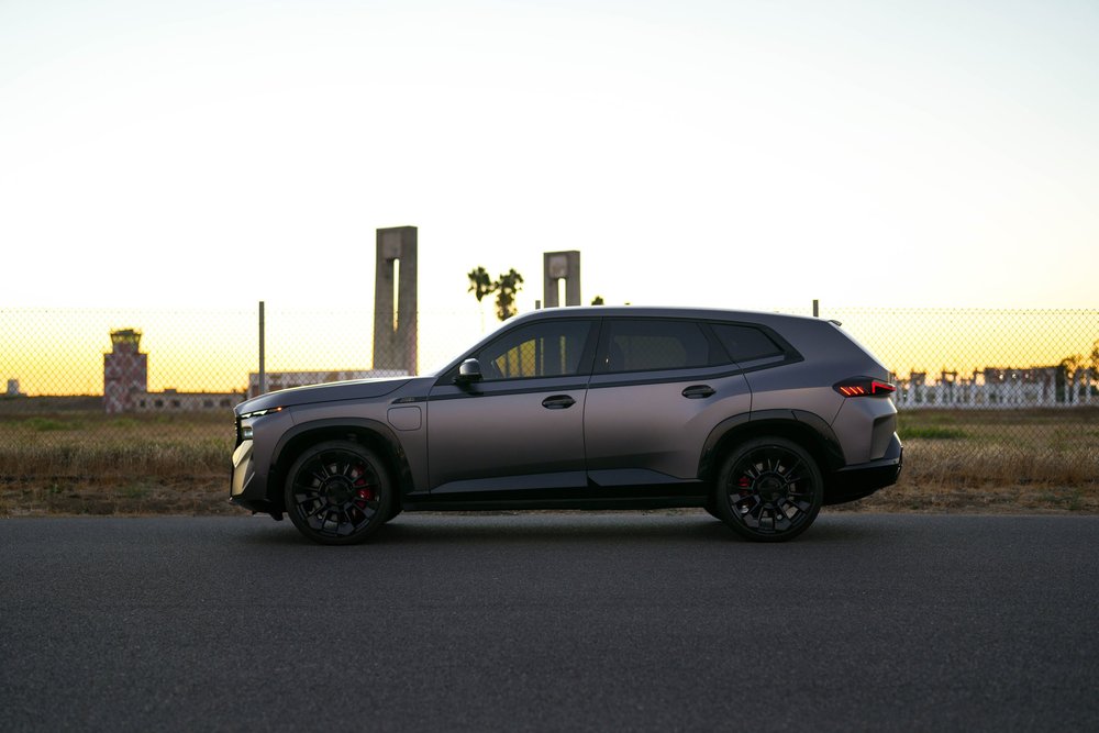 Complete side profile of the matte-grey BMW XM with dark windows and black wheels against the sunset and roadside fence.