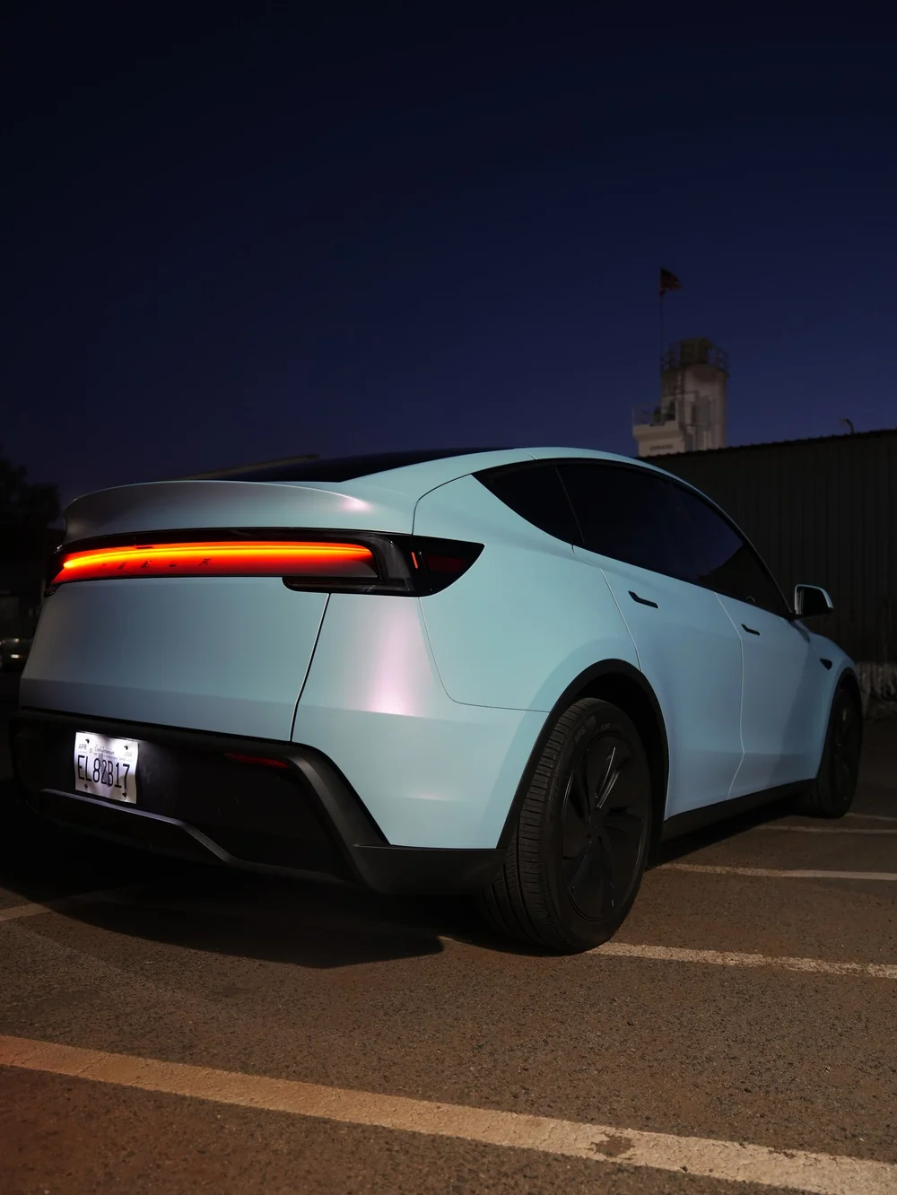 Rear three-quarter view of a Tesla Model Y with white-to-blue vinyl under outdoor lighting