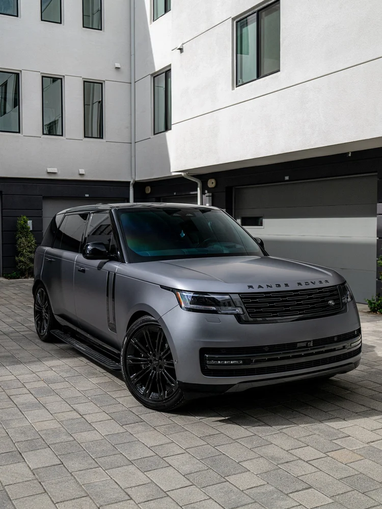 Complete front three-quarter view of a satin grey Range Rover LWB with gloss black wheels on paving beside a light building
