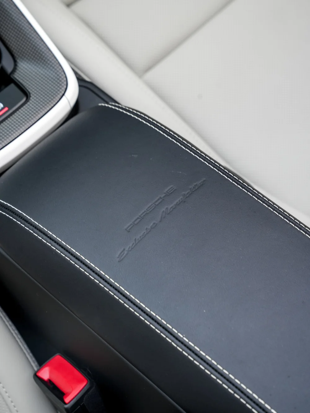 Dark stitched Porsche center armrest beside the light seat and red belt latch
